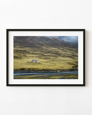 An abandoned Icelandic farmhouse beside a river beneath grassy hills fading into low mist.
