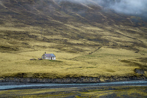 A lone white farmhouse with a weathered metal roof in a broad, misty Icelandic valley.