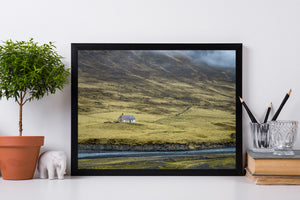 Remote Icelandic house above a blue river, surrounded by rolling yellow-green hills.
