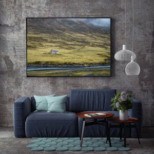 Weathered rural house beside a river and steep grass-covered valley slopes in Iceland.