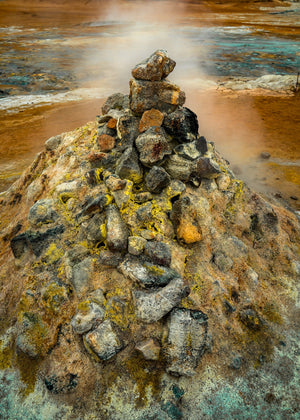 Stacked dark rocks above yellow deposits and ochre ground in a steaming Icelandic geothermal field.