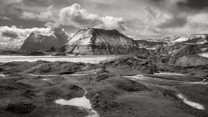 Steep Icelandic mountain slopes rise beyond dark uneven terrain and small reflective pools.
