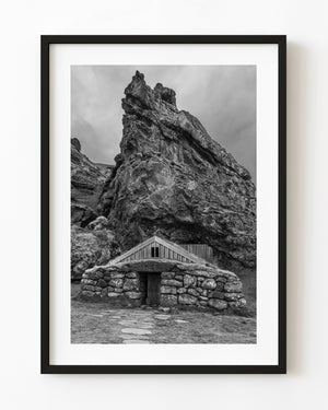 Icelandic turf house with stone walls beneath a towering rock formation, photographed in black and white.
