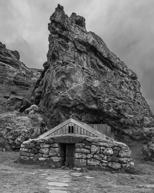 A small turf-roofed house and wooden gable sheltered beneath a rugged Icelandic rock face.