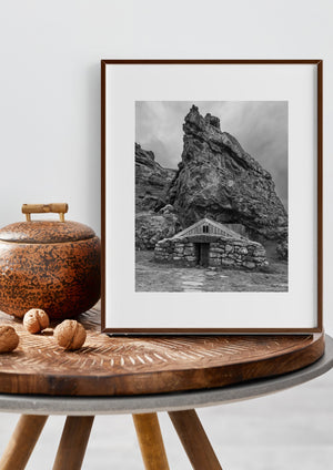 Turf-covered stone house dwarfed by a jagged rock face in a monochrome Iceland landscape.