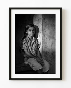Black and white portrait of an Indian girl seated on a windowsill beside a sunlit textured wall.