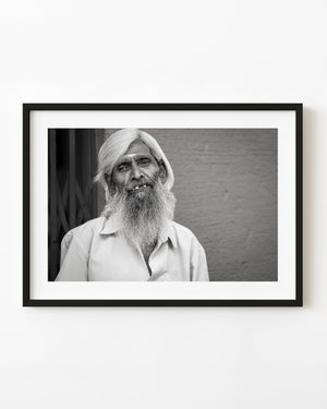 Black and white portrait of an Indian man with a long white beard smiling beside a textured wall.