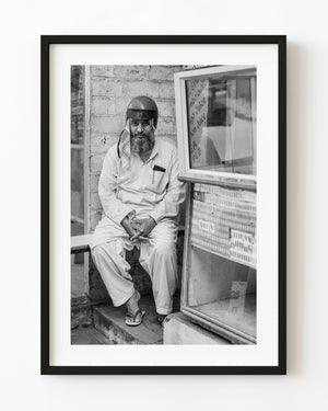 Black and white portrait of an Indian man in a motorcycle helmet seated beside a shop display.