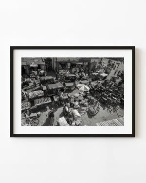 Black and white view from above of an Indian street market filled with produce carts, people and vehicles.