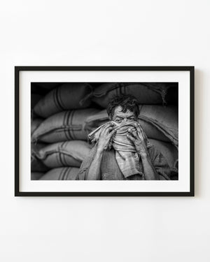 Black and white portrait of an Indian worker adjusting a scarf across his face beside stacked sacks.