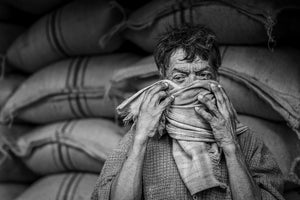 An Indian man holds a woven scarf over his nose and mouth against a backdrop of sacks.