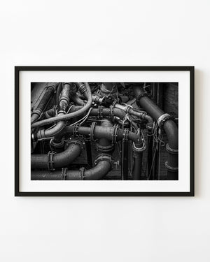 Black and white close-up of industrial iron pipes, bolted joints and valves forming a dense mechanical pattern.