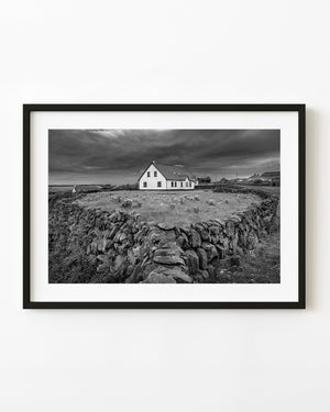 Black and white Irish cottage behind a curving stone wall beneath a dramatic cloudy sky.