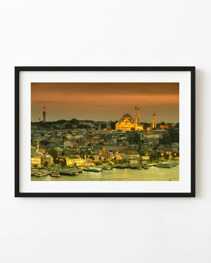 Istanbul waterfront beneath a golden sunset sky, with illuminated domes, minarets and boats along the shore.