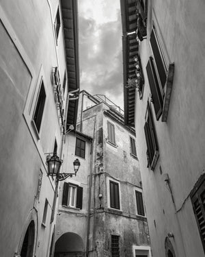 Tall Italian buildings frame a narrow strip of cloudy sky above an alley in monochrome.
