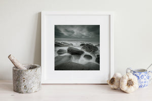 Long-exposure photograph of Kostbergan Beach with dark stone and pale surf.