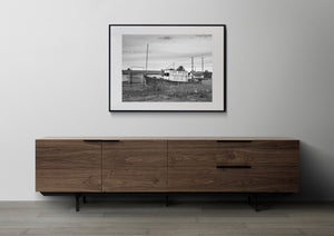 Monochrome rural scene with an abandoned charter boat and small wooden buildings.