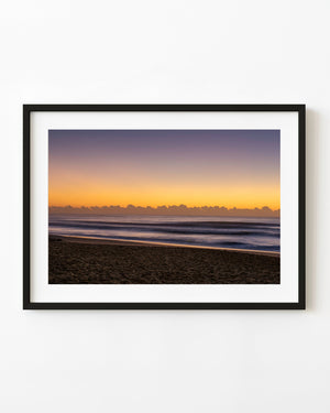 Long Reef Beach at sunrise with dark sand, soft waves and a warm golden sky
