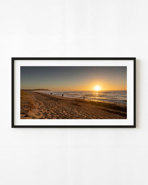 People walking along Long Reef Beach beneath a golden sunrise over the sea