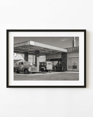 Vintage trucks beneath a weathered Ludlow service station canopy in black and white