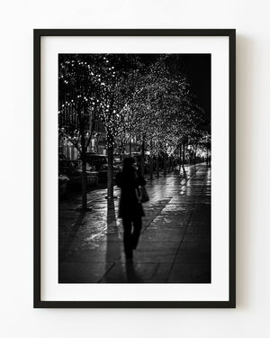 Black and white photograph of a woman walking on a wet Manhattan sidewalk at night