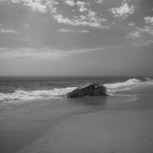 A dark beach boulder rests at the water’s edge beneath scattered clouds