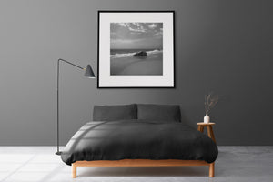 Square black and white beach photograph with a boulder, open sea and pale sky