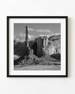 Black and white photograph of a slender sandstone spire and cliffs in Monument Valley