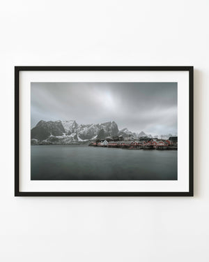 Red Norwegian waterfront cabins beneath snow-covered mountains and a grey sky