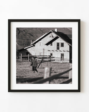 Romanian farmer carrying a shovel across a fenced field in black and white