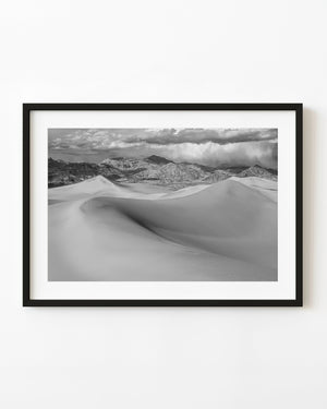 Sand dunes and rugged mountains beneath storm clouds in black and white
