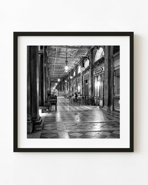 Black and white cafe arcade at night in St Marks Square, Venice, with glowing lamps and patterned paving.