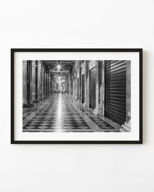 Black and white Venice night arcade with repeating columns, closed shutters and diamond-patterned paving.