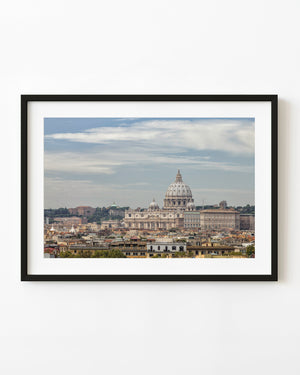 St Peters Basilica rises above Rome rooftops beneath a pale blue clouded sky.