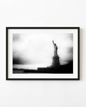 Statue of Liberty above a dark island shoreline beneath a bright clouded sky in black and white.