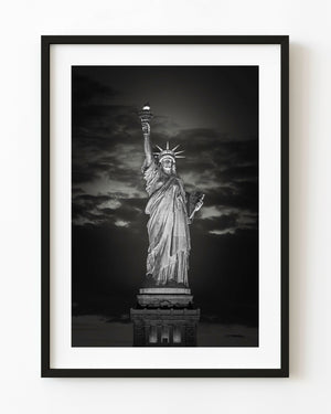 Illuminated Statue of Liberty at night against dark clouds in black and white.