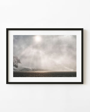 Sunlight filters through an Antarctic snowstorm above dark water and snowy coastal mountains.