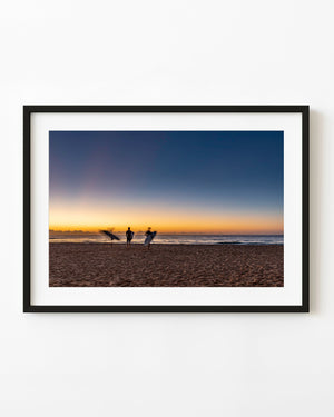 Surfers carrying boards at sunrise on Long Reef Beach, Sydney, beneath a blue and gold sky.