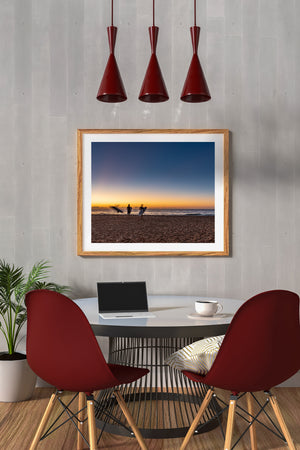 Warm dawn light glows above the ocean behind surfers and textured sand.
