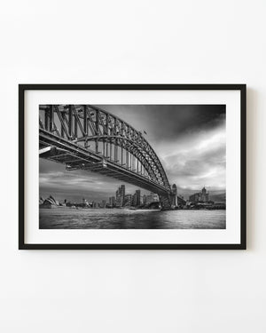 Sydney Harbour Bridge beneath dramatic clouds, with the city skyline and Opera House in black and white.
