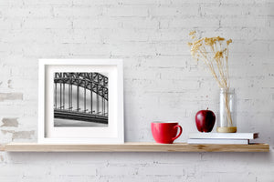 Parallel bridge supports divide pale sky in a square black and white photograph.