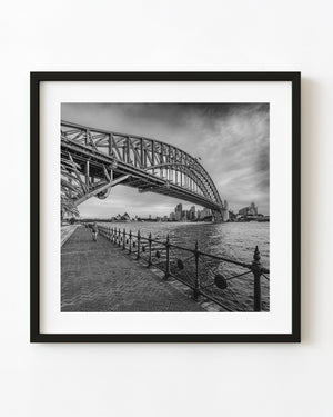 Sydney Harbour Bridge above a waterfront walk, with the Opera House and skyline in black and white.