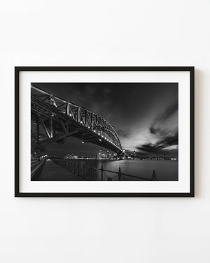 Sydney Harbour Bridge illuminated at night above calm water and the city skyline in black and white.