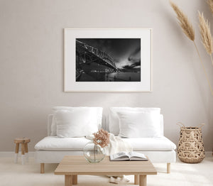 Waterfront railing leads beneath Sydney Harbour Bridge in a black and white night photograph.