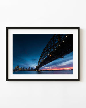 Sydney Harbour Bridge viewed from below at sunset, with blue sky and an orange harbour horizon.