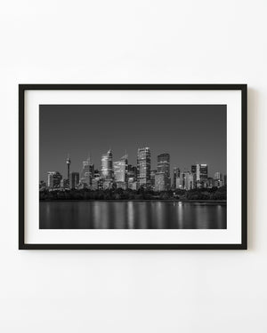 Sydney skyline lights reflected in smooth harbour water in a black and white night photograph.