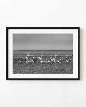 Three white Adirondack chairs overlook the St Lawrence shoreline in black and white.