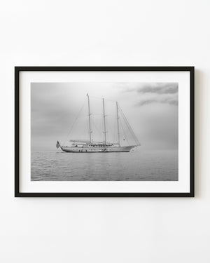 Three-masted sailing yacht rests on calm water beneath a pale cloudy sky in black and white.