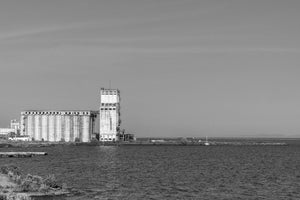 Weathered concrete silos and a tall elevator tower stand beneath a broad pale sky.