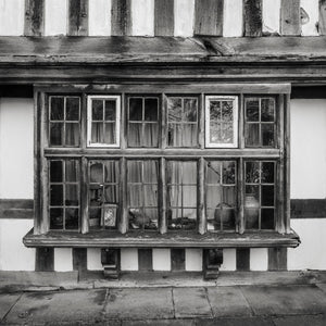 Weathered wooden beams surround an old English window with curtains and small objects inside.